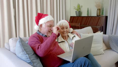 Pareja-Caucásica-Mayor-Con-Sombreros-De-Santa-Usando-Una-Computadora-Portátil-Para-Videollamadas-Navideñas,-Cámara-Lenta