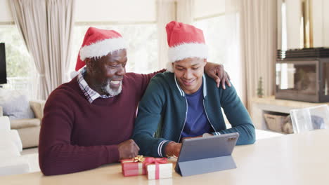 Happy-african-american-father-and-adult-son-in-christmas-hats-having-tablet-video-call,-slow-motion