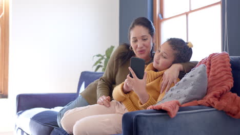 Feliz-Madre-E-Hija-Birraciales-Abrazándose-En-El-Sofá-Y-Usando-Un-Teléfono-Inteligente-En-Una-Soleada-Sala-De-Estar