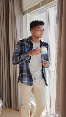 Vertical-video-of-african-american-man-drinking-coffee-looking-out-of-sunny-window,-slow-motion