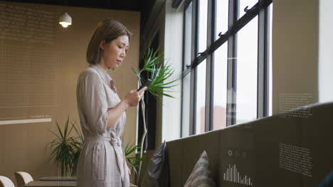 Animation-of-graphs,-loading-bars-and-circles,-asian-woman-standing-near-window-and-using-cellphone