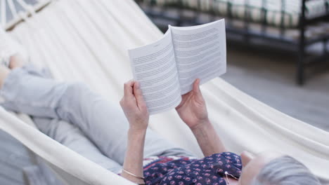 Caucasian-middle-aged-woman-reading-book-relaxing-in-hammock-on-terrace-in-nature,-slow-motion