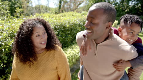 Happy-african-american-mother-and-father-carrying-son-on-back-walking-in-sunny-garden