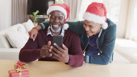 Feliz-Padre-E-Hijo-Afroamericanos-Con-Sombreros-Navideños-Teniendo-Videollamadas-Con-Teléfonos-Inteligentes,-Cámara-Lenta