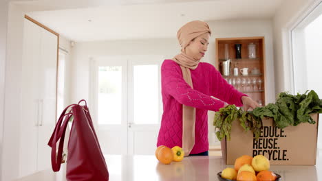 Feliz-Mujer-Birracial-En-Hijab-Con-Compras-En-La-Cocina-De-Casa-Con-Espacio-Para-Copiar,-Cámara-Lenta