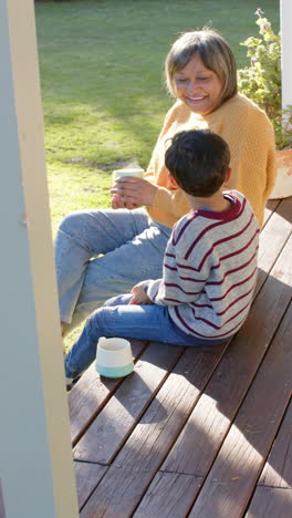 Vertical-video-of-biracial-grandmother-and-grandson-sitting-on-terrace-holding-mugs,-slow-motion
