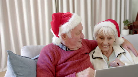 Pareja-Caucásica-Mayor-Con-Sombreros-De-Santa-Usando-Una-Computadora-Portátil-Para-Videollamadas-Navideñas,-Cámara-Lenta