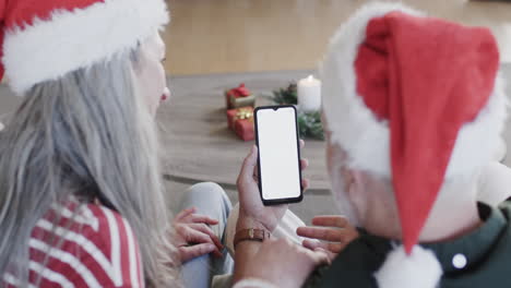 Pareja-Caucásica-Con-Sombreros-De-Santa-Smartphone-Con-Pantalla-En-Blanco-En-Navidad-En-Casa,-Cámara-Lenta