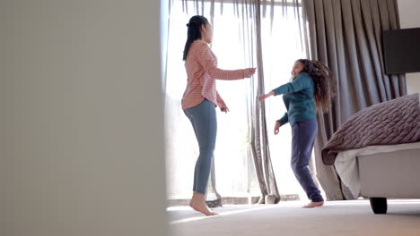 Happy-biracial-mother-and-daughter-dancing-in-sunny-bedroom