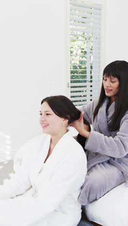 Vertical-video-of-happy-biracial-mother-brushing-hair-of-adult-daughter-in-sunny-room,-slow-motion