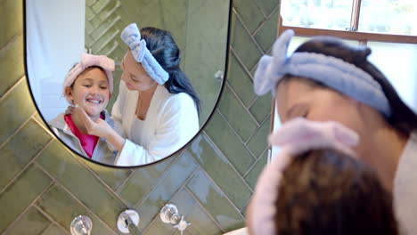 Happy-biracial-mother-and-daughter-with-headbands-taking-care-of-faces-in-sunny-bathroom