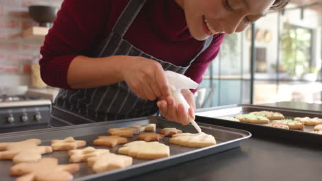 Feliz-Mujer-Birracial-En-Delantal-Decorando-Galletas-Navideñas-En-La-Cocina,-Cámara-Lenta