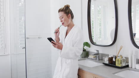 Mujer-Birracial-Cepillándose-Los-Dientes-Usando-Un-Teléfono-Inteligente-En-El-Baño,-Cámara-Lenta