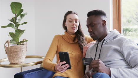 Happy-diverse-couple-sitting-on-sofa,-using-smartphone-and-credit-card-in-home,copy-space