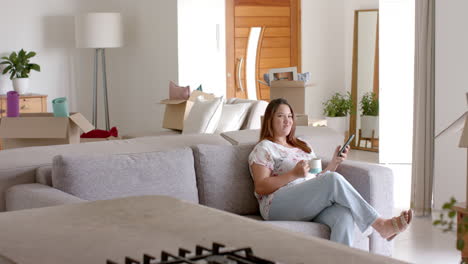 Mujer-Birracial-De-Talla-Grande-En-Un-Sofá-Usando-Un-Teléfono-Inteligente-Tomando-Café-En-Un-Nuevo-Hogar,-Espacio-Para-Copiar,-Cámara-Lenta