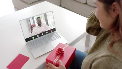 Caucasian-woman-holding-red-gift-using-laptop-with-african-american-man-on-screen