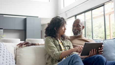 Happy-biracial-couple-sitting-on-couch-and-using-tablet-at-home,-slow-motion