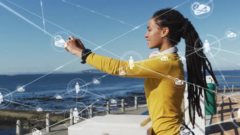 Animation-of-network-of-connections-with-icons-over-biracial-woman-using-smartphone-by-sea