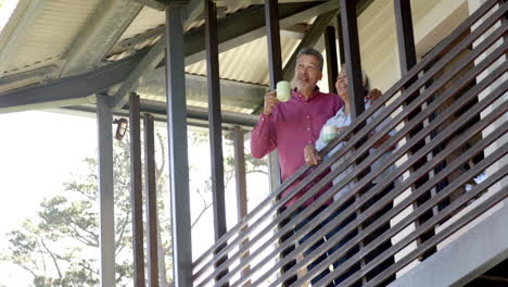 Happy-biracial-senior-couple-embracing-on-balcony,-talking-and-drinking-tea