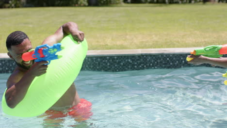 Felices-Padres-Afroamericanos-E-Hija-Jugando-Con-Pistolas-De-Agua-En-Una-Piscina-Soleada,-Cámara-Lenta