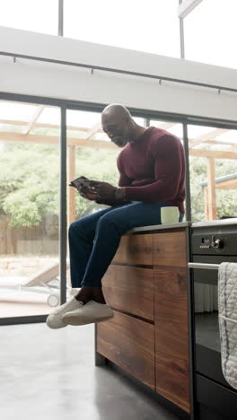 Video-Vertical-De-Un-Hombre-Afroamericano-Usando-Un-Teléfono-Inteligente-Y-Celebrando-En-La-Cocina,-Cámara-Lenta