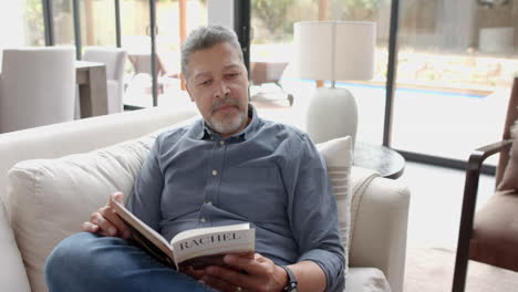 Hombre-Birracial-Senior-Enfocado-Sentado-En-El-Sofá-Leyendo-Un-Libro-En-La-Sala-De-Estar-En-Casa,-Cámara-Lenta