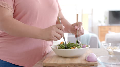 Feliz-Mujer-Birracial-De-Talla-Grande-Lanzando-Un-Tazón-De-Ensalada-De-Verduras-Feta-En-La-Cocina,-Cámara-Lenta