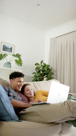 Vertical-video-of-happy-diverse-couple-sitting-on-sofa-using-laptop-at-home,-in-slow-motion