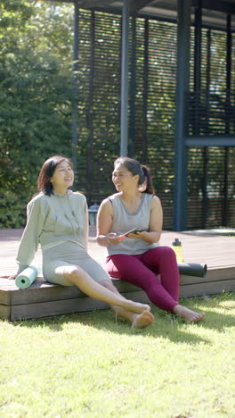 Felices-Amigas-Asiáticas-Con-Colchonetas-De-Ejercicio-Usando-Un-Teléfono-Inteligente-En-Una-Terraza-Soleada,-Cámara-Lenta