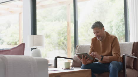 Happy-senior-biracial-man-using-tablet-sitting-on-sofa-in-sunny-living-room,-copy-space,-slow-motion