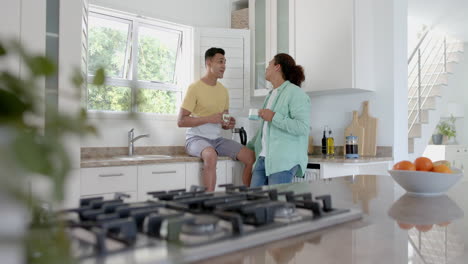 Happy-diverse-gay-male-couple-having-coffee-and-talking-in-sunny-kitchen,-copy-space,-slow-motion