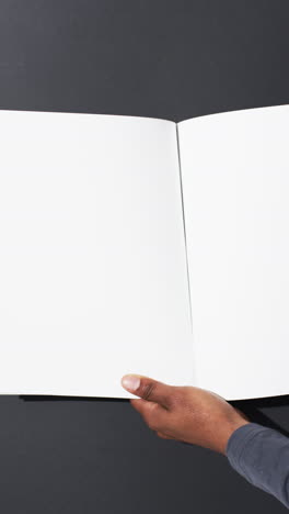 Vertical-video-of-hand-of-african-american-man-with-book-with-white-blank-pages-on-grey-background