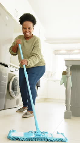 Happy-african-american-senior-woman-cleaning-floor-in-sunny-kitchen,-slow-motion