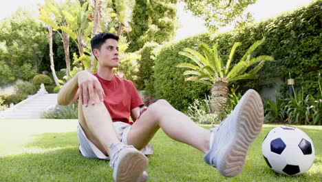 Focused-biracial-man-sitting-with-football,-touching-toes-in-sunny-garden,-copy-space,-slow-motion