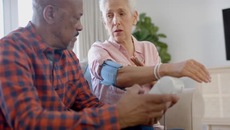 Seria-Pareja-Birracial-Senior-Sentada-En-El-Sofá-Y-Controlando-La-Presión-Arterial-En-Casa,-Cámara-Lenta