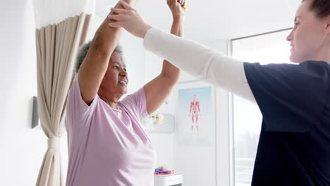 Diverse-senior-woman-exercising-with-band-and-female-doctor-advising-in-hospital,-slow-motion