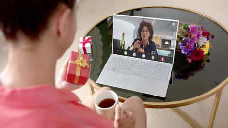 Mujer-Caucásica-Sosteniendo-Un-Regalo-Rojo-Usando-Una-Computadora-Portátil-Con-Un-Hombre-Birracial-En-La-Pantalla