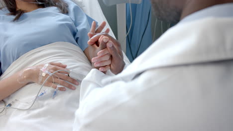 Diverse-male-doctor-holding-hand-of-female-patient-with-oximeter-in-hospital-bed,-slow-motion