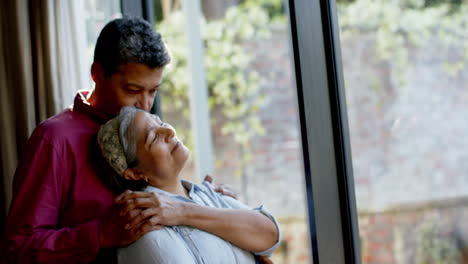 Happy-biracial-senior-couple-embracing-at-window-at-home