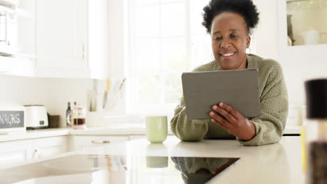 Feliz-Mujer-Mayor-Afroamericana-Tomando-Café,-Usando-Una-Tableta-En-Una-Cocina-Soleada,-Cámara-Lenta