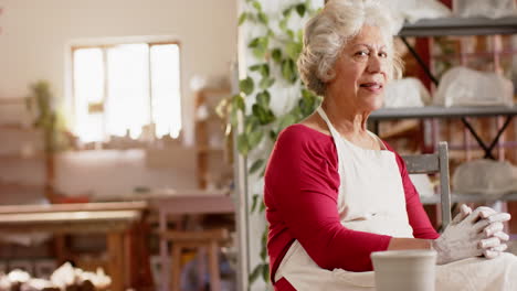 Happy-senior-biracial-female-potter-with-gray-hair-resting-smiling-in-pottery-studio,-slow-motion