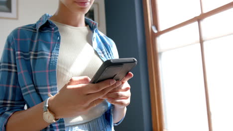 Focused-biracial-teenage-girl-standing-in-sunny-room-using-smartphone,-slow-motion