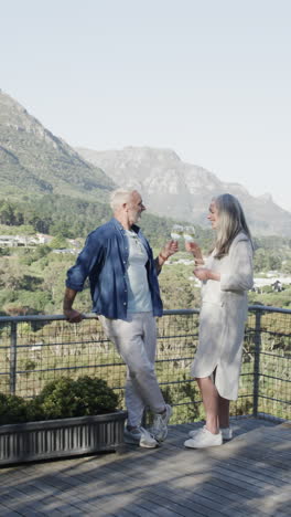 Vertical-video-of-happy-senior-caucasian-couple-standing-on-balcony-drinking-wine,-slow-motion