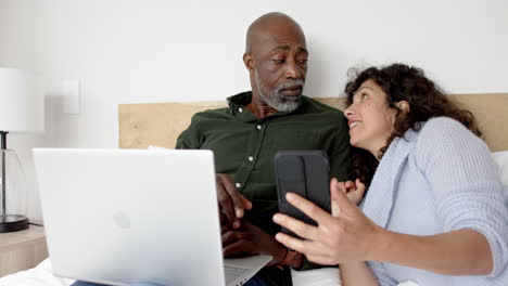 Happy-mature-diverse-couple-sitting-on-bed-using-laptop-and-smartphone-at-home,-slow-motion