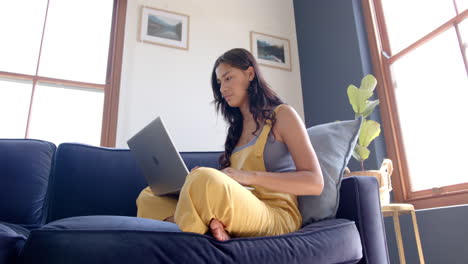 Focused-biracial-teenage-girl-sitting-on-couch-using-laptop,-smiling,-copy-space,-slow-motion