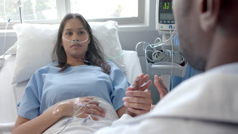 Diverse-male-doctor-holding-hand-of-female-patient-with-oximeter-in-hospital-bed,-slow-motion