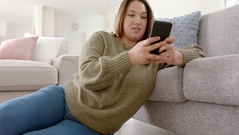 Happy-plus-size-biracial-woman-relaxing-in-living-room-using-smartphone,-slow-motion