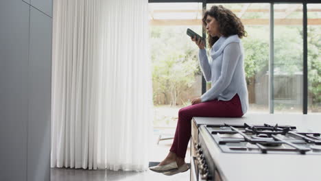 Happy-mature-caucasian-woman-sitting-on-kitchen-counter-and-talking-on-smartphone,-slow-motion