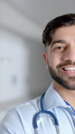 Vertical-video-of-portrait-of-happy-biracial-male-doctor-in-hospital-corridor,-slow-motion