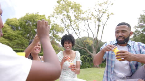 Happy-african-american-parents,-daughter-and-son-blowing-bubbles-sitting-in-garden,-slow-motion